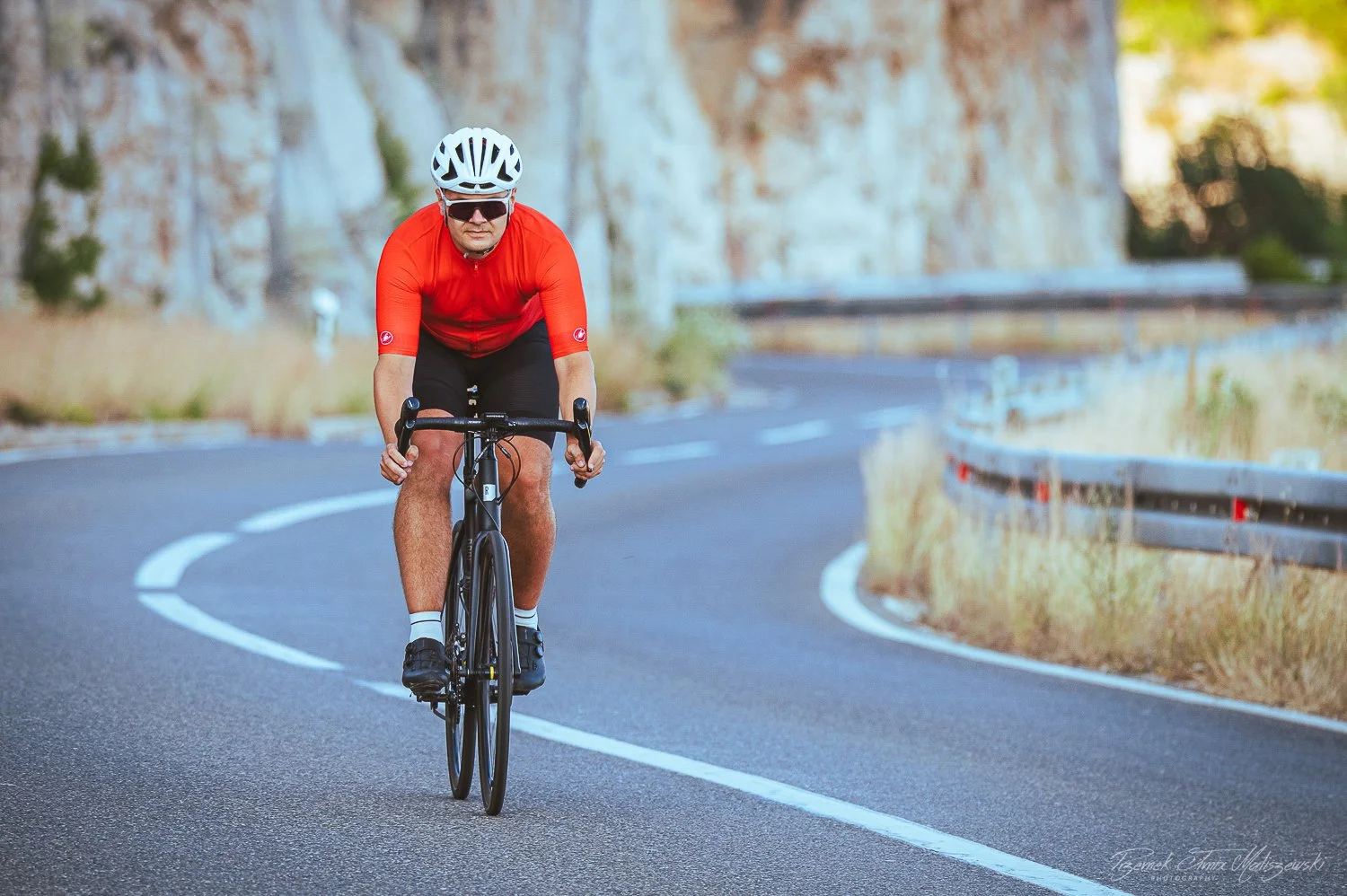 A man wearing a red shirt, black shorts, a white helmet, and sunglasses riding a black road bike on a winding mountain road lined with guardrails and dry grass.