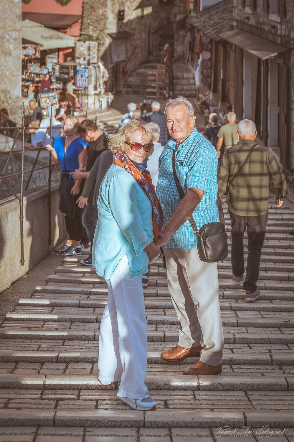 An elderly couple holding hands and looking at the camera on a stone staircase in a busy, historic outdoor market or town area with other people in the background.