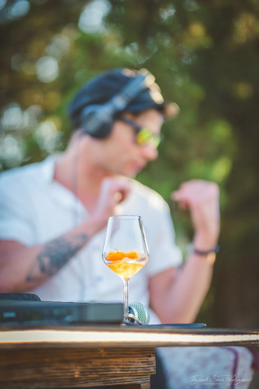 A person wearing a cap, sunglasses, and headphones, making a fist pump gesture, with a wine glass with orange drink and fruit slices on a wooden table in the foreground, outdoors with blurred trees and sunlight in the background.