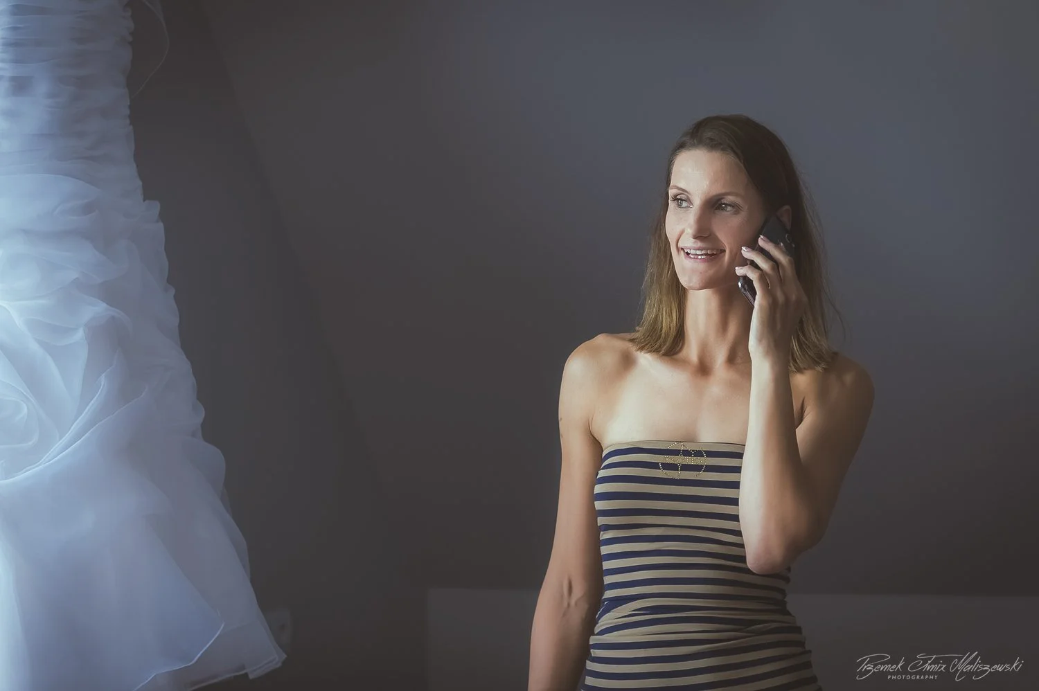 Young woman with shoulder-length brown hair wearing a strapless navy and beige striped dress, smiling and talking on her cellphone in a dimly lit room.