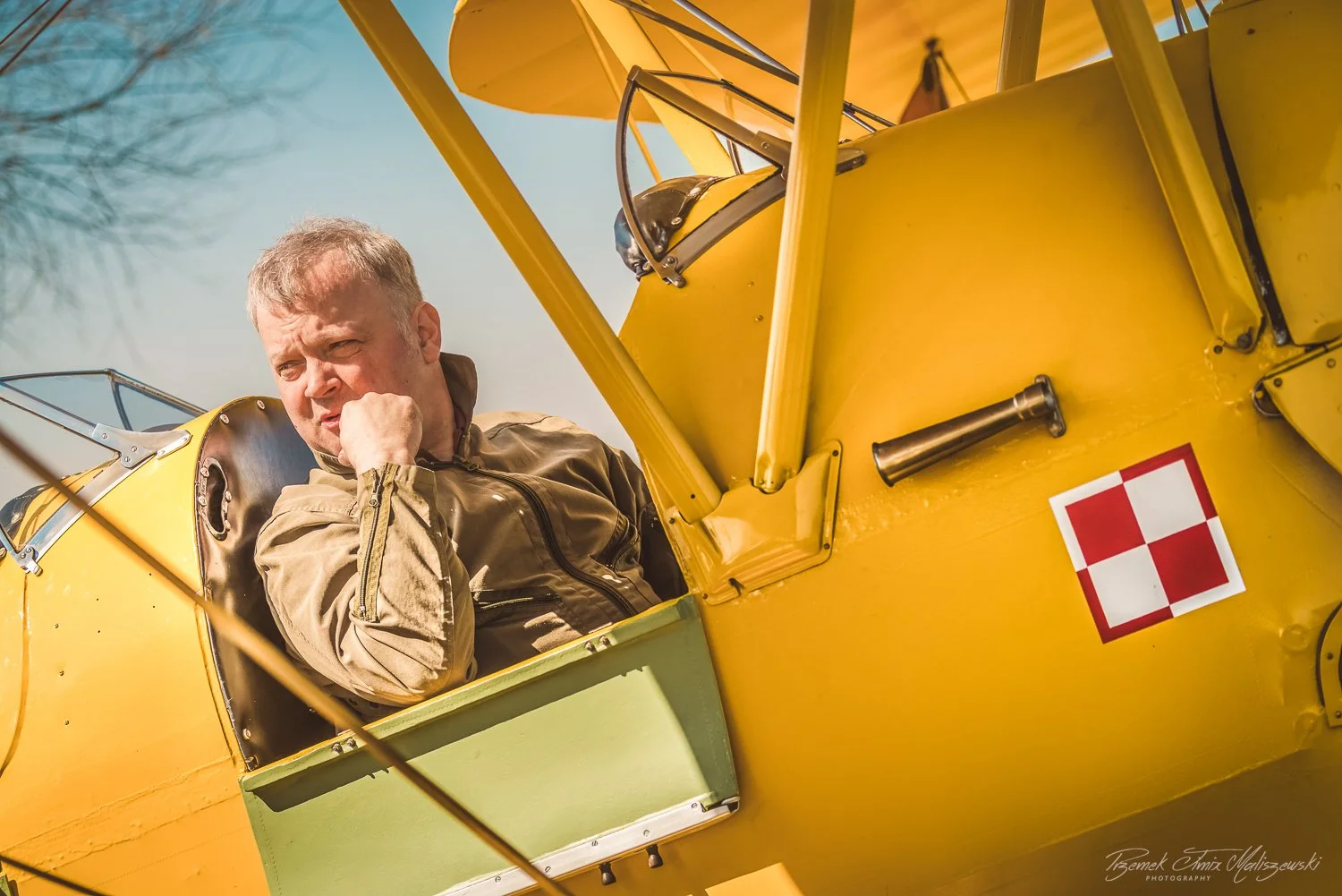 A man sitting in the cockpit of a vintage yellow airplane, wearing a beige flight jacket, looking concerned with his chin resting on his fist.