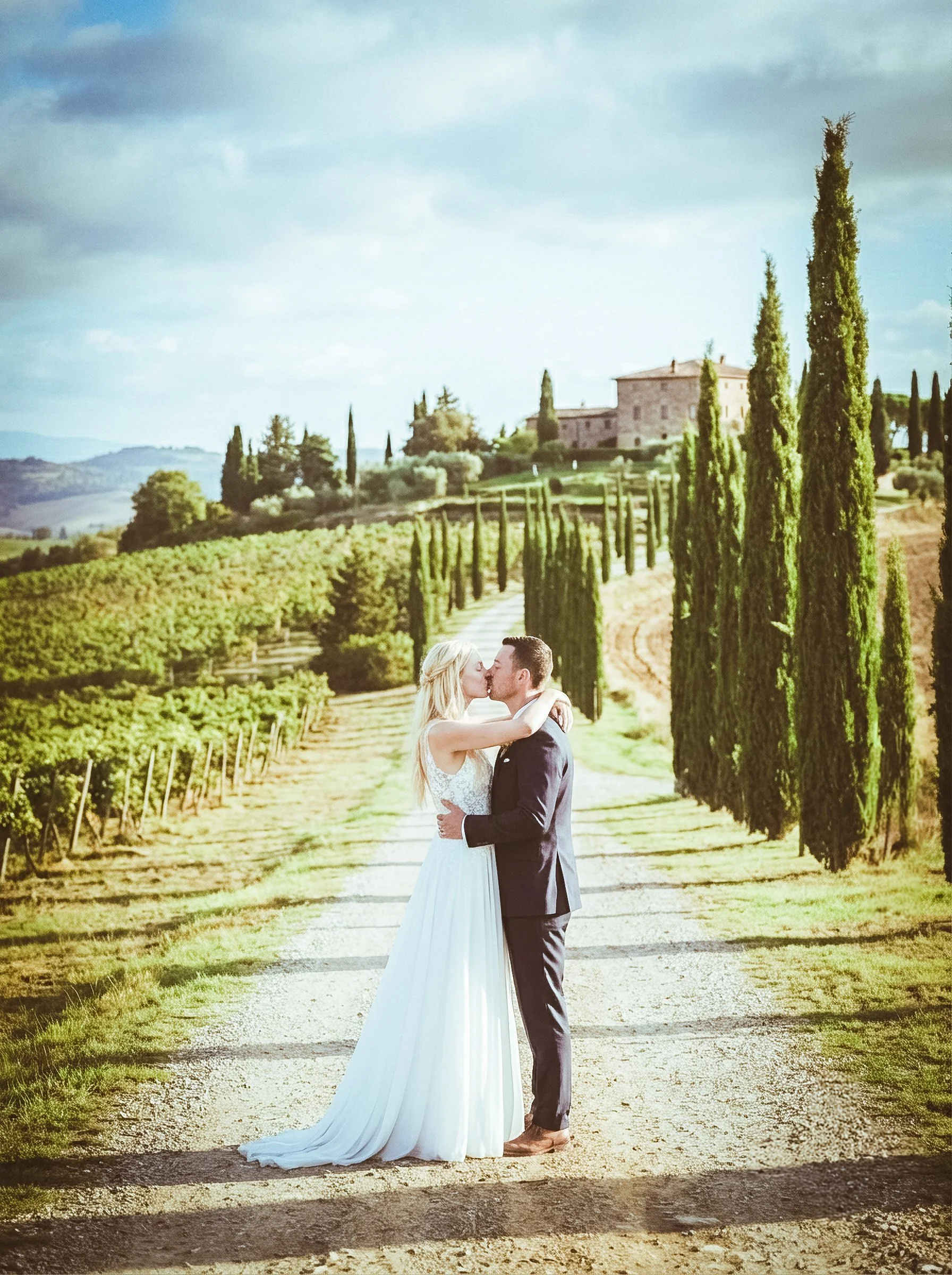 A bride and groom kissing in a vineyard with a large estate in the background, tall cypress trees lining the path, and rolling hills in the distance