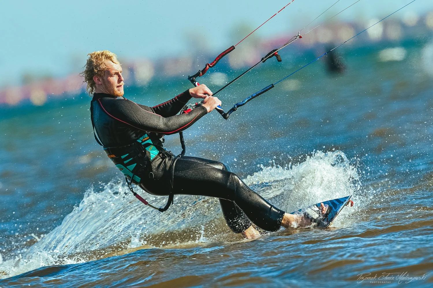 A man kite surfing on the water, wearing a black wetsuit, holding the kite's control bar with a focused expression, with water splashing around him.