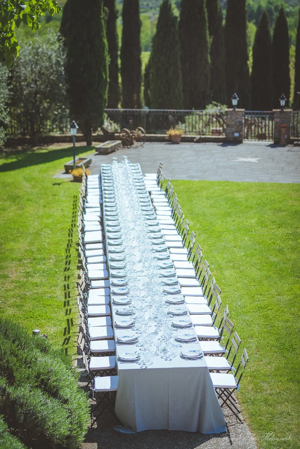 Long dining table set outdoors with white tablecloth, plates, and glassware on a green lawn, surrounded by trees and a stone patio.