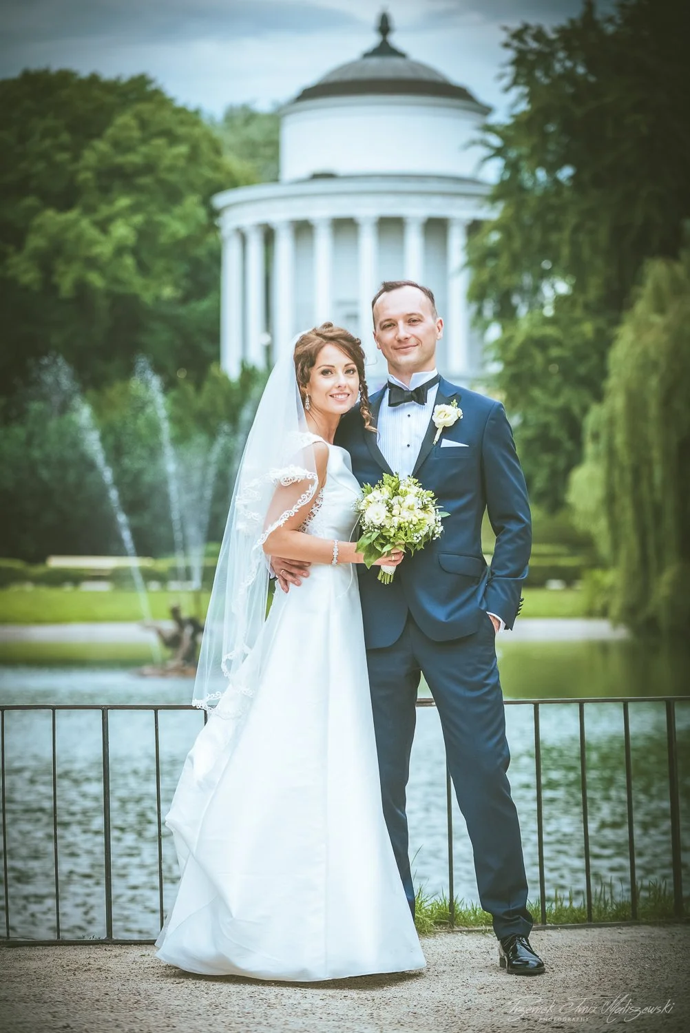 A bride and groom standing by a lake with a fountain and a white, round building with columns in the background, during their wedding photoshoot.