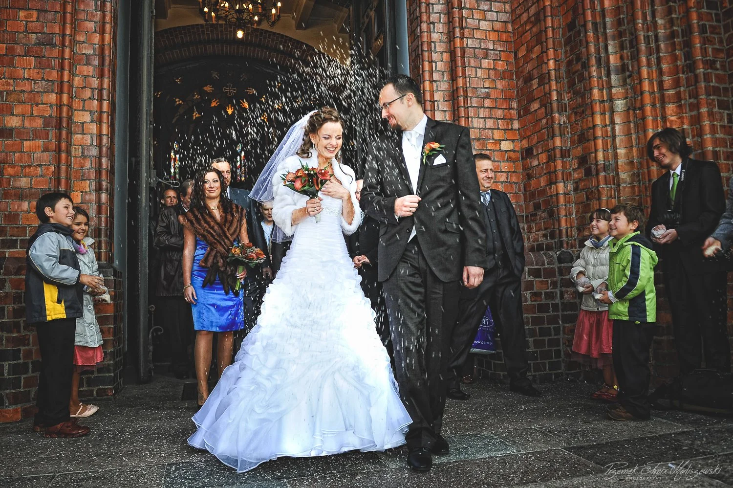 A bride and groom celebrating their wedding outside a brick church, being splashed with water by guests.