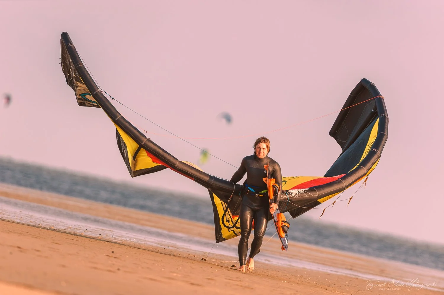 A man in a wetsuit standing barefoot on the beach, holding a kiteboarding kite with a large inflatable wing, with the ocean and other kite surfers in the background.