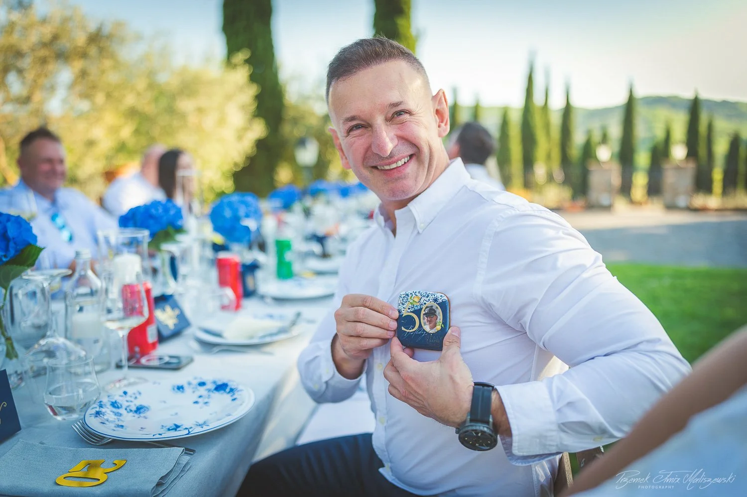 A smiling man in a white shirt holding a decorated box with a photo at an outdoor celebration table with blue flowers and place settings.