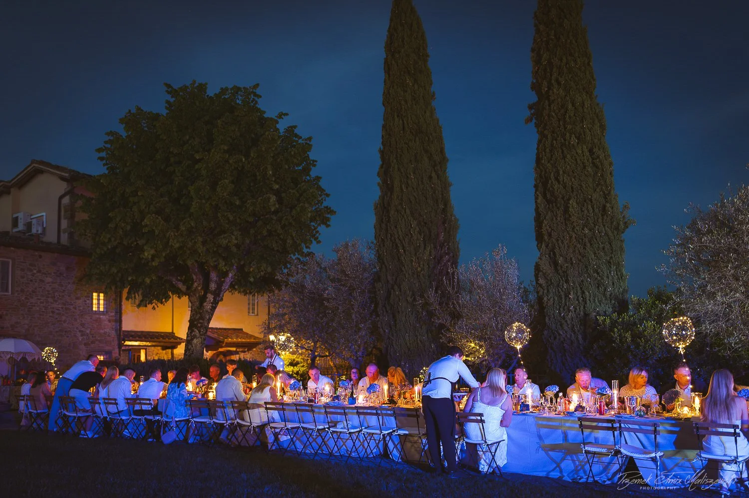 A large outdoor dinner party at night with people sitting at long tables decorated with candles and flowers, surrounded by tall trees and a stone building, with hanging string lights and round illuminated decorations.