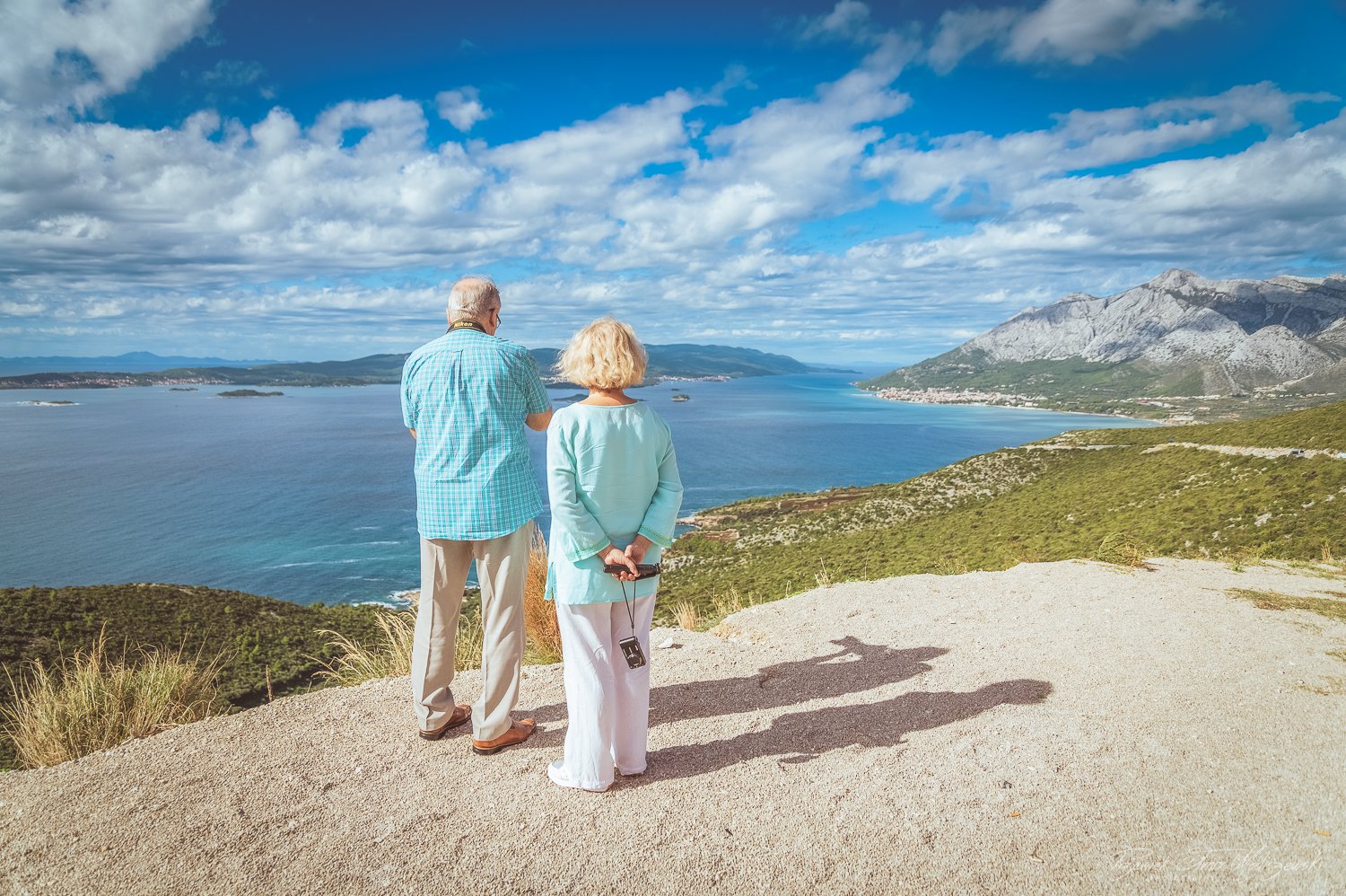 An elderly man and woman standing on a viewing platform overlooking a scenic bay with mountains in the distance under a partly cloudy sky.