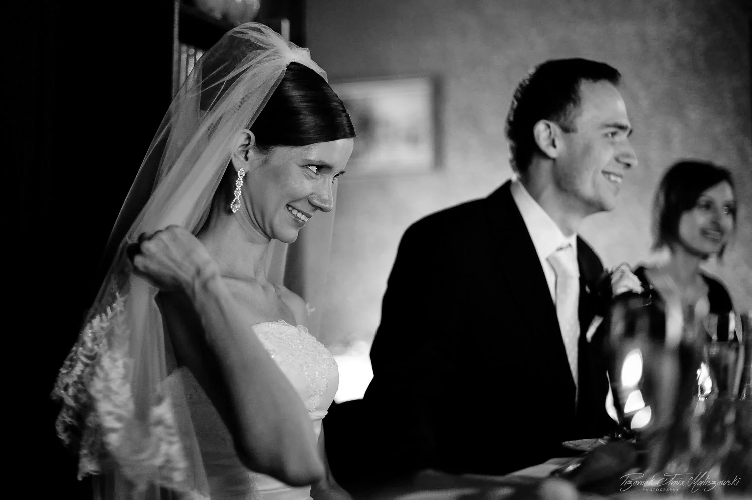Black and white photo of a bride and groom at their wedding reception. The bride is smiling, adjusting her veil, and wearing elegant earrings. The groom is also smiling, dressed in a suit, and sitting next to her. In the background, a woman with shor