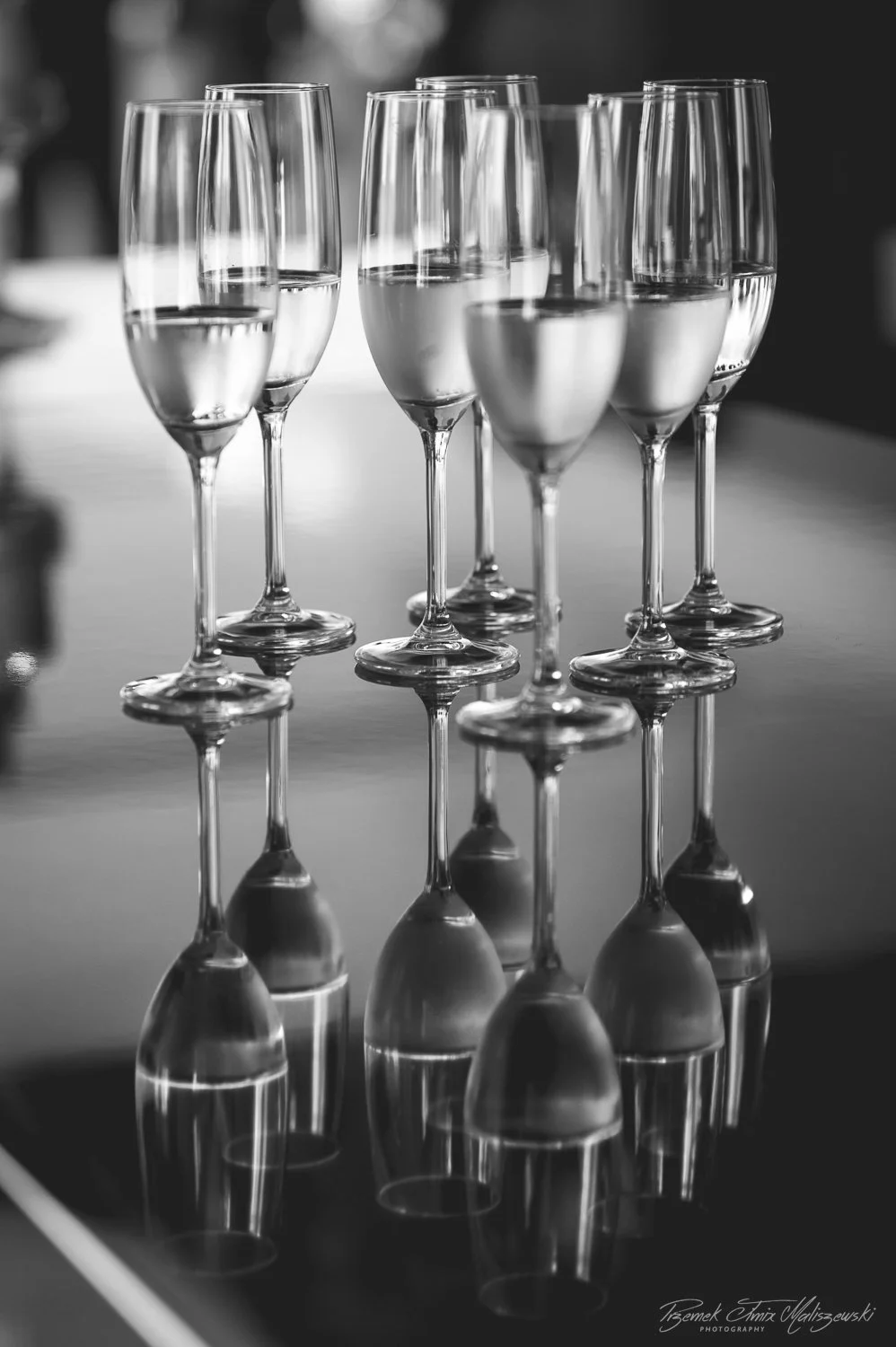 Six champagne flutes filled with a clear liquid, arranged on a reflective surface, with the reflection visible beneath them.