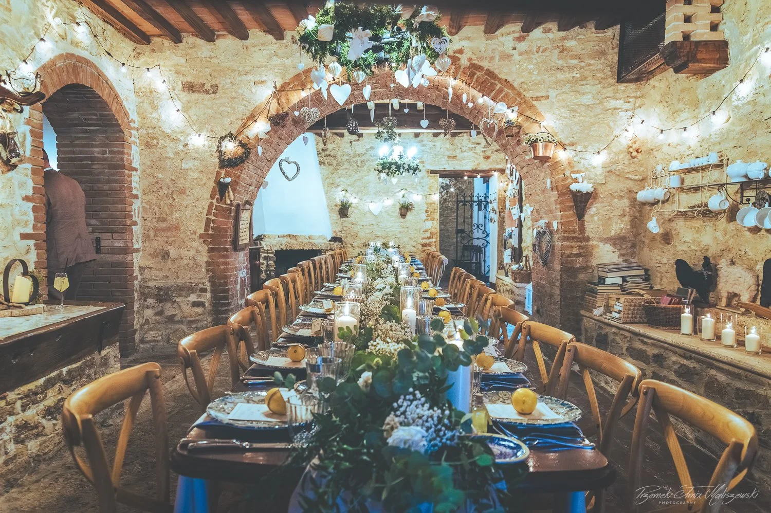 A long dining table set for a celebration inside a rustic stone and brick-walled room decorated with hanging heart-shaped ornaments, string lights, and candles.