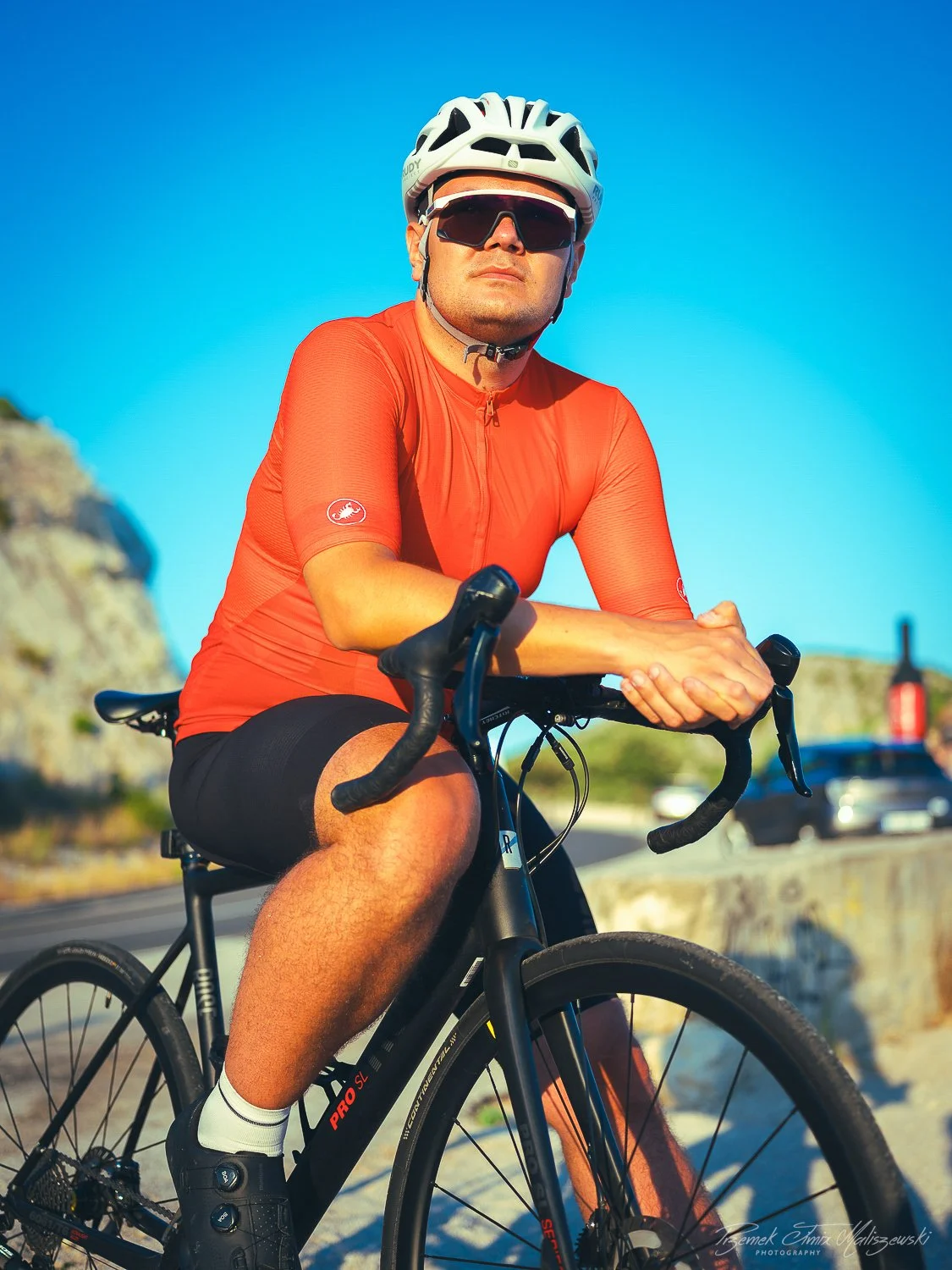A man wearing sunglasses, a white helmet, and a red cycling jersey riding a black road bike outdoors during the daytime.