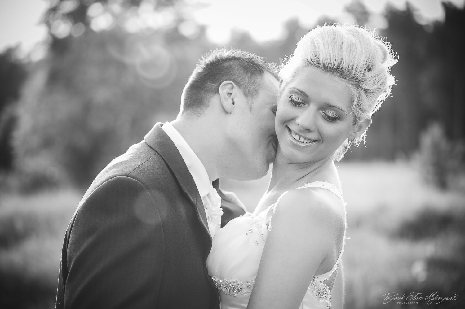 Black and white photo of a man in a suit kissing a smiling woman in a sleeveless dress outdoors, with trees in the background.