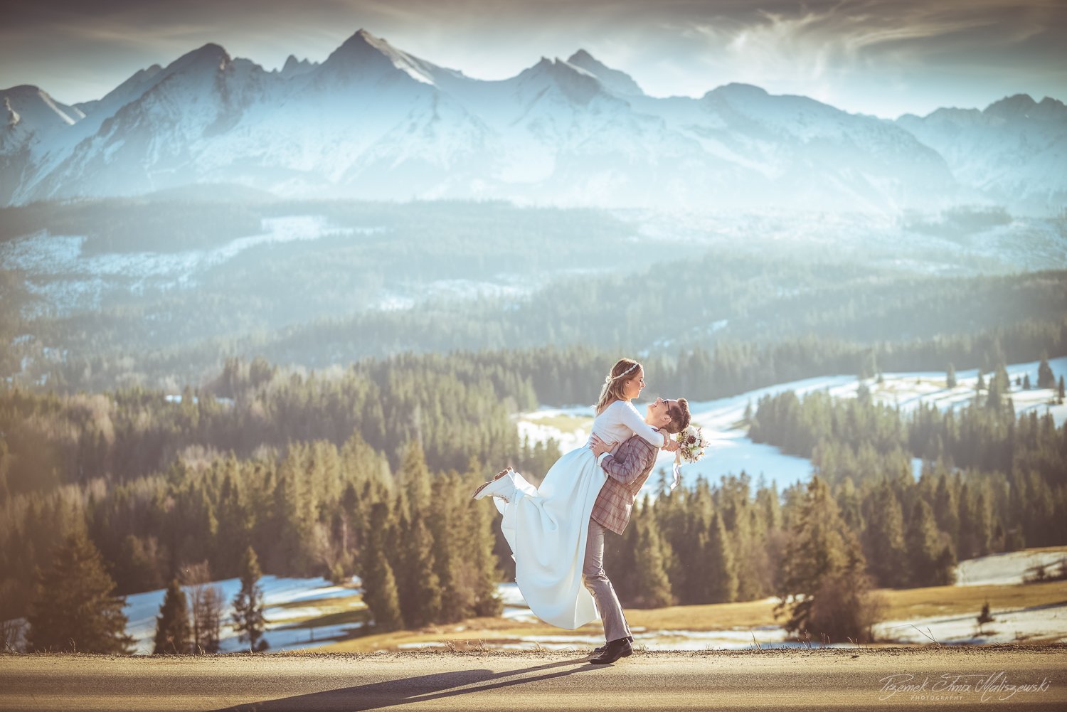 A man lifting a woman in a white dress in a scenic mountainous landscape with snow-capped peaks and dense forests.
