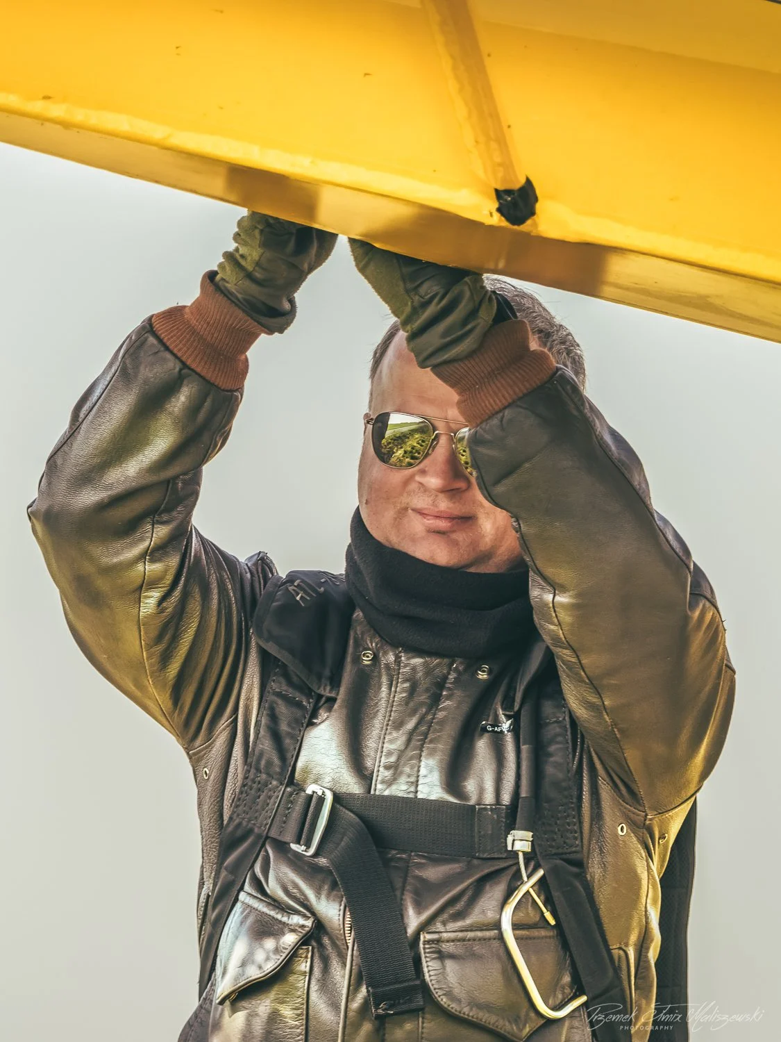 A man wearing sunglasses, a leather jacket, and a black neck gaiter holding an object above his head. The man is smiling slightly and looking towards the camera.