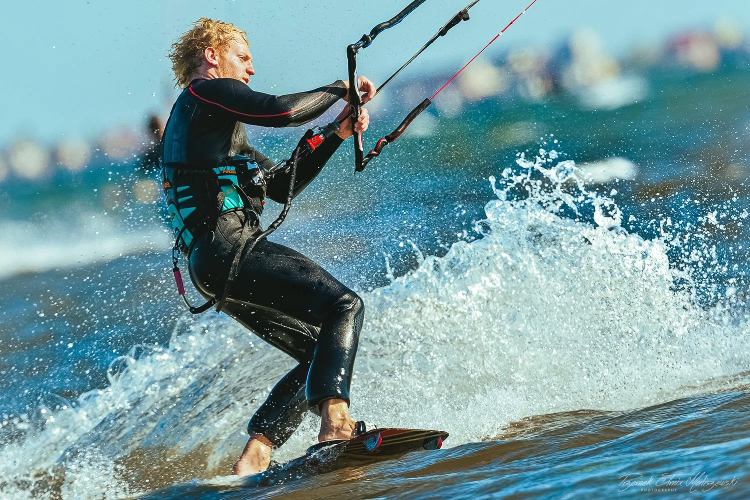 A person kite surfing on the water, holding onto a control bar with both hands, with a sail attached to their harness, wearing a wetsuit, and leaning back as the surfboard moves through the water
