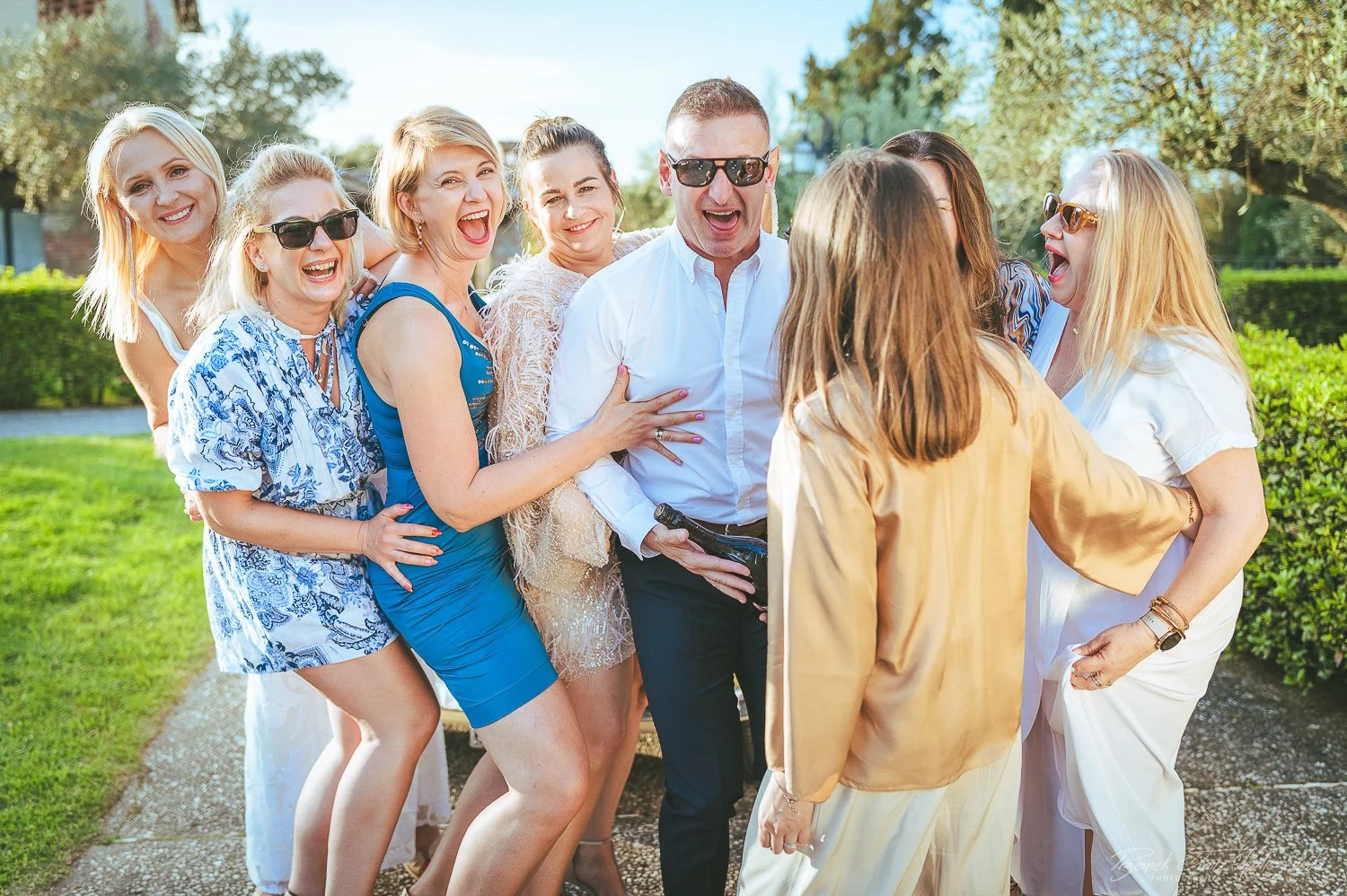 A group of women and a man are gathered outdoors, smiling and laughing, wearing casual summer clothing, with trees and greenery in the background.