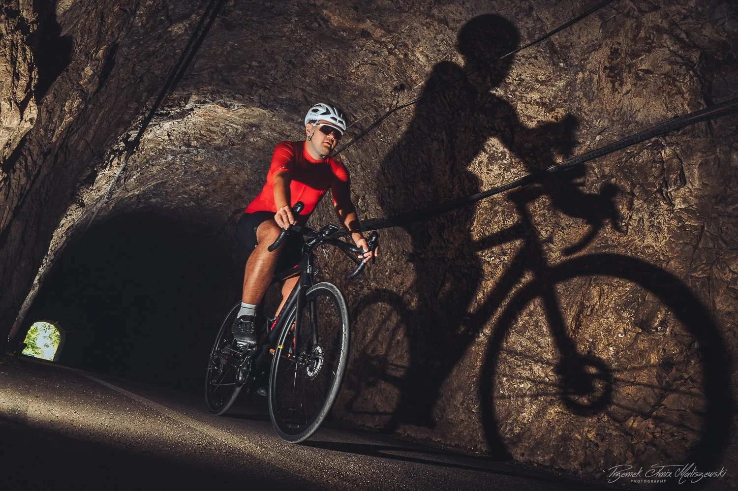 A cyclist in a red shirt and helmet riding a bicycle through a rocky tunnel, with the shadow of the cyclist and bike cast on the tunnel wall.