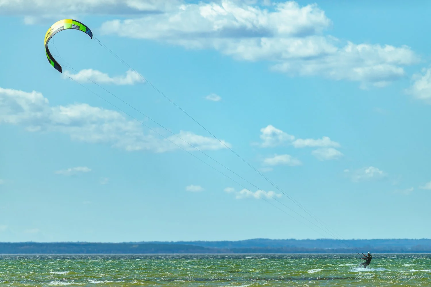 A person kiteboarding on the water with a large green and black kite in the sky, with a distant landmass and a partly cloudy sky in the background.