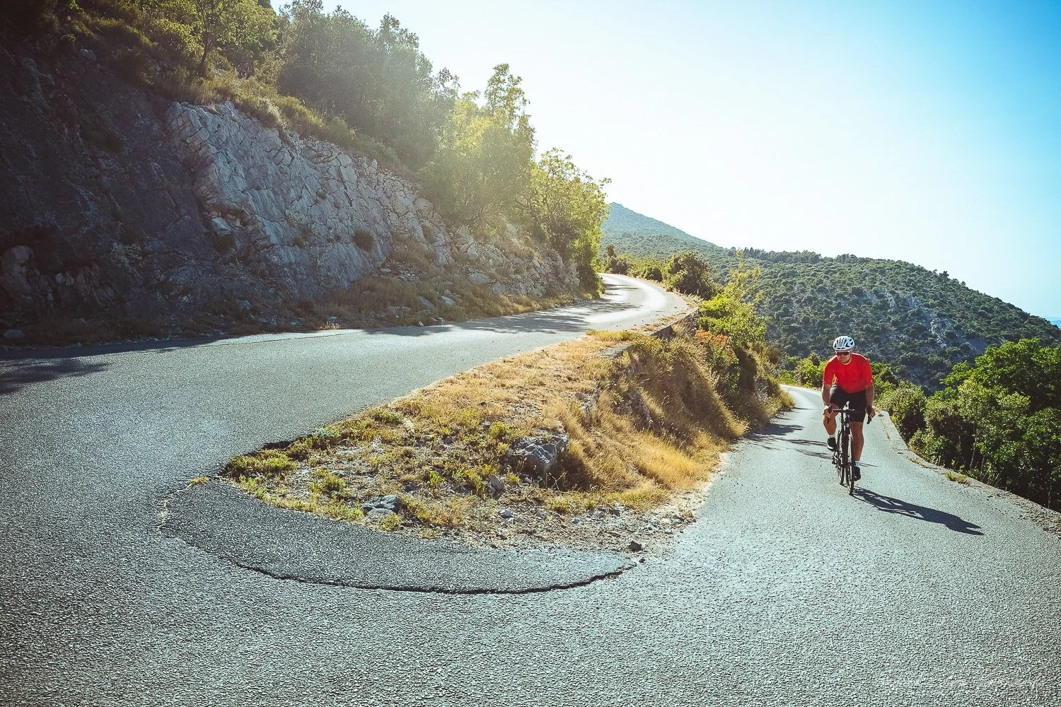 A cyclist wearing a red shirt and helmet riding a bicycle on a winding mountain road surrounded by hills with sparse vegetation and trees, under a clear blue sky.