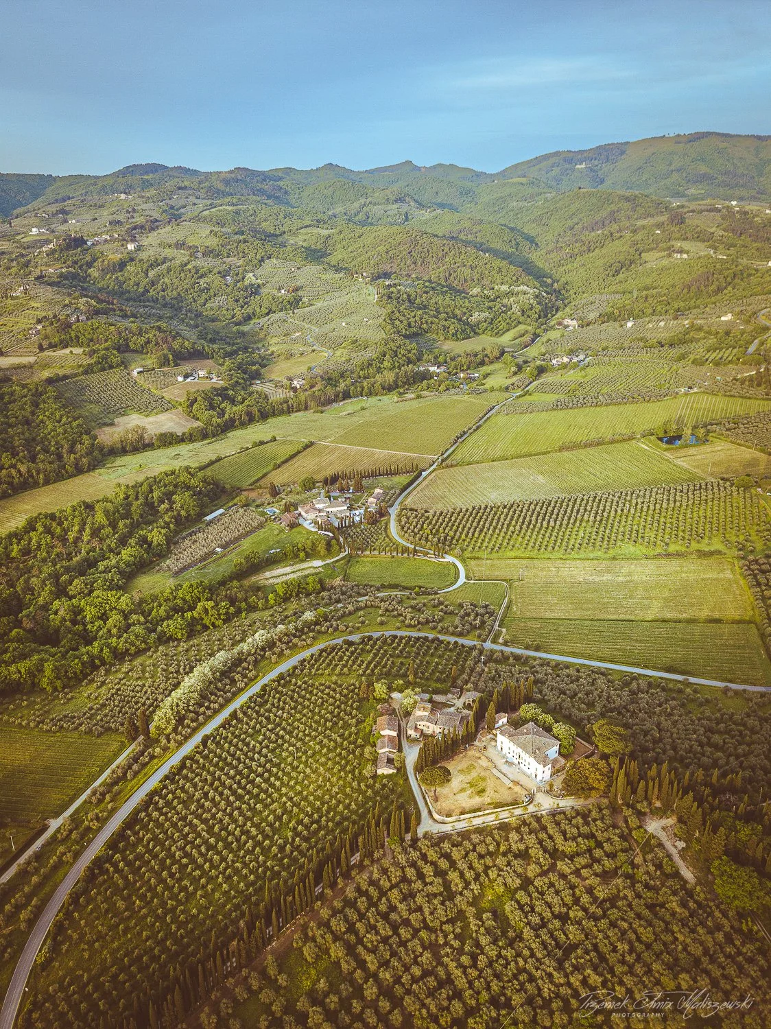 Aerial view of lush green rolling hills with farmland, winding roads, and scattered buildings in a rural landscape.