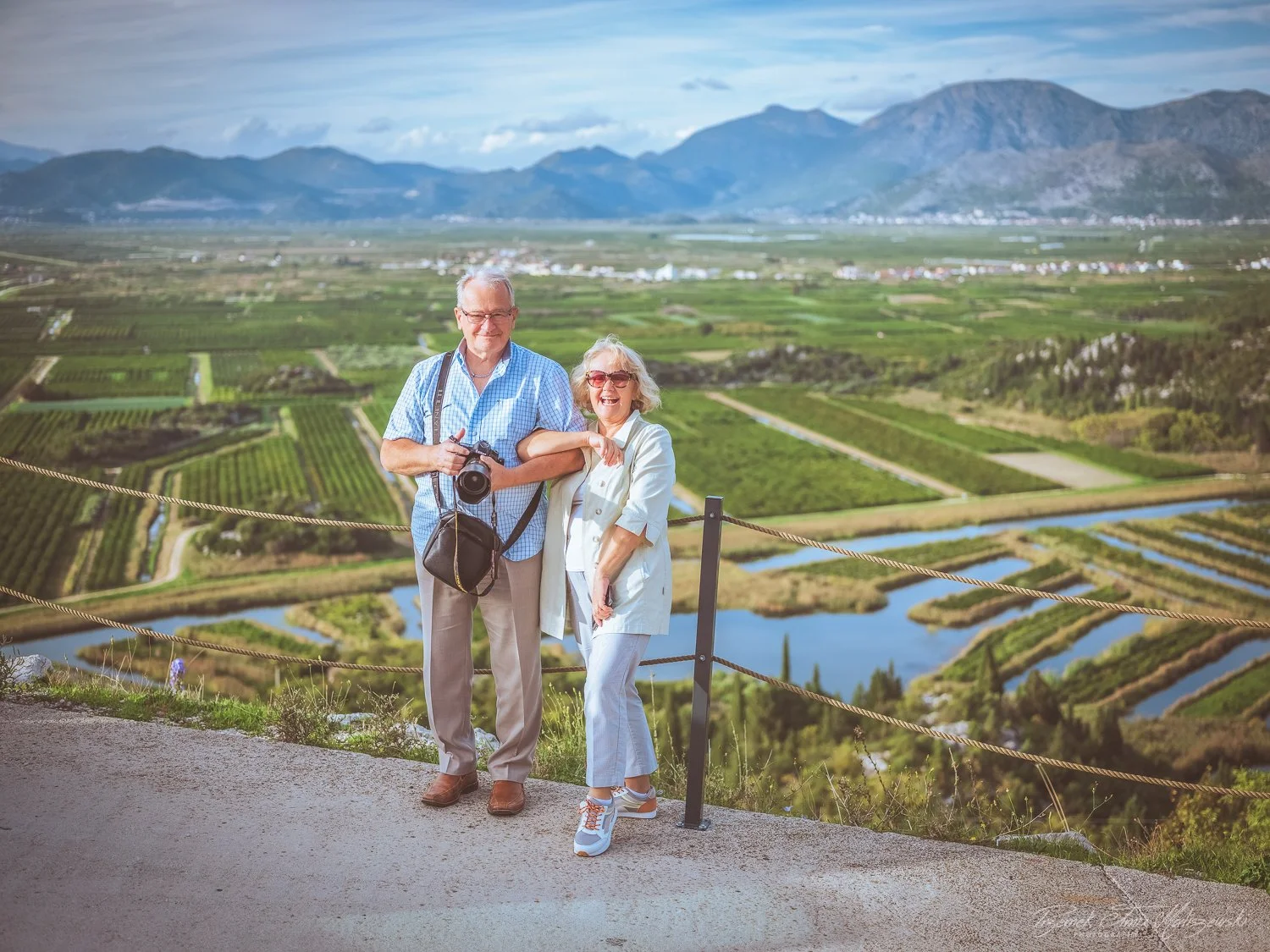 An elderly couple standing on a scenic overlook with a view of farmlands, water channels, and mountains in the background