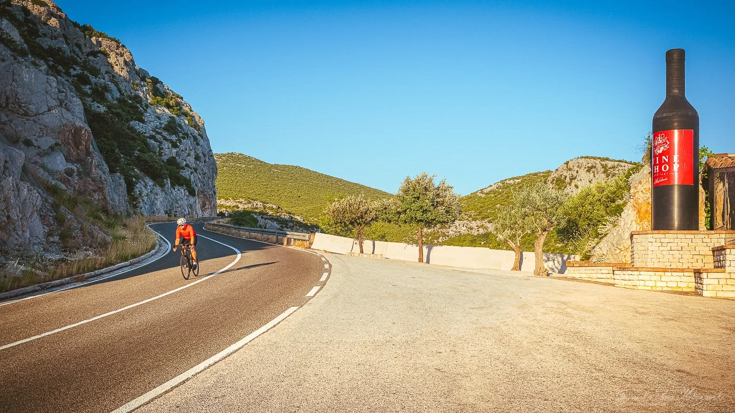 A cyclist wearing a red shirt and helmet riding on a winding mountain road with a large bottle-shaped structure with a red label on the side.