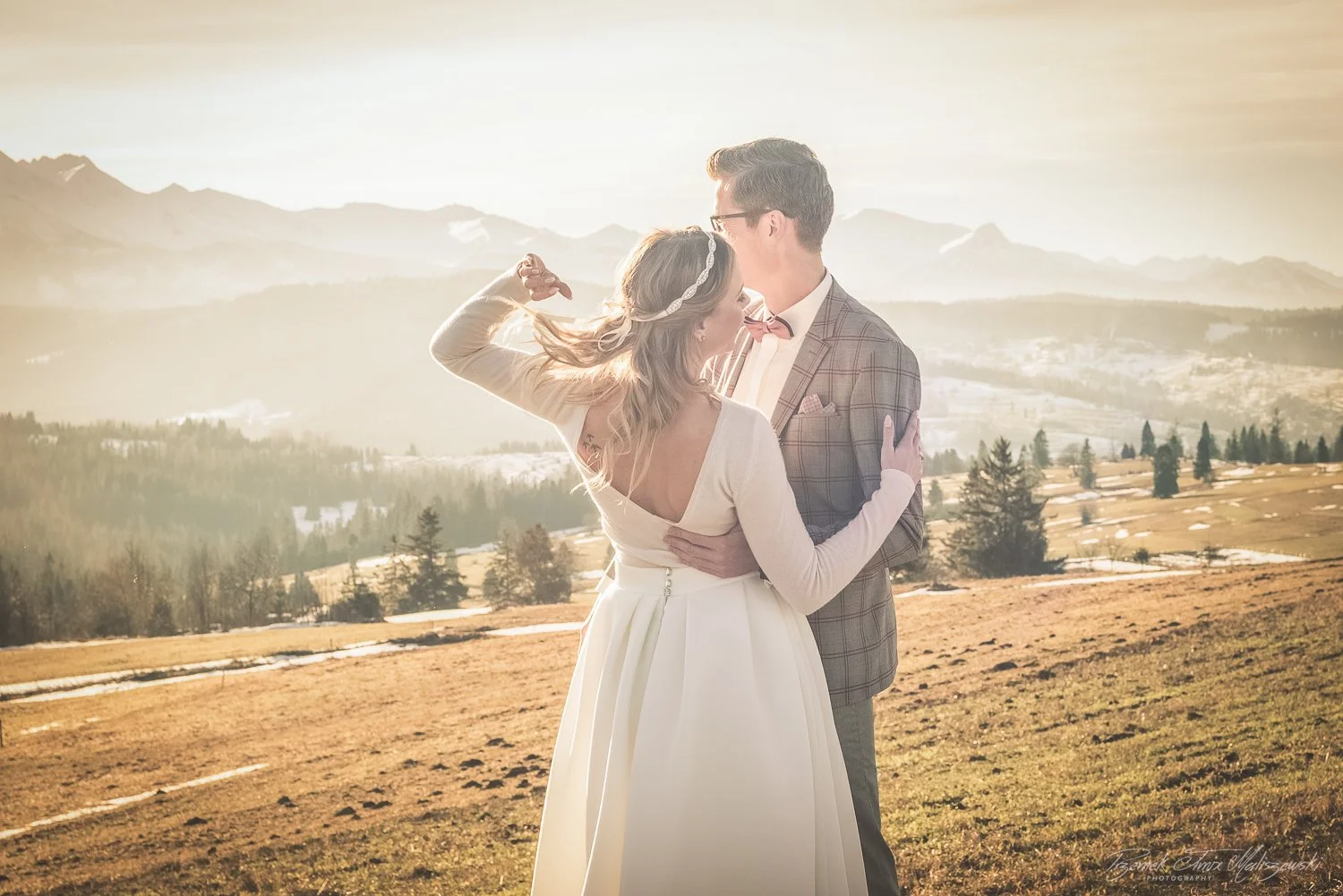 A bride and groom embracing outdoors in a mountainous landscape during sunset.
