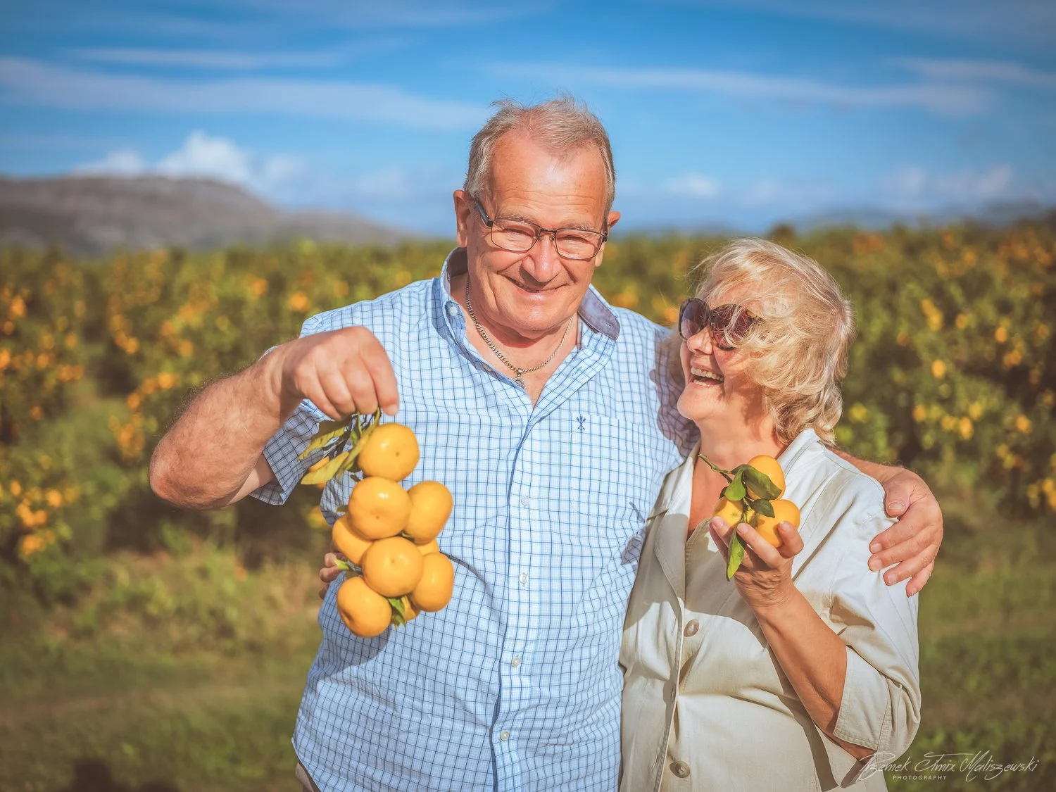 An elderly couple smiling and holding ripe yellow citrus fruit in a lush orchard on a sunny day.