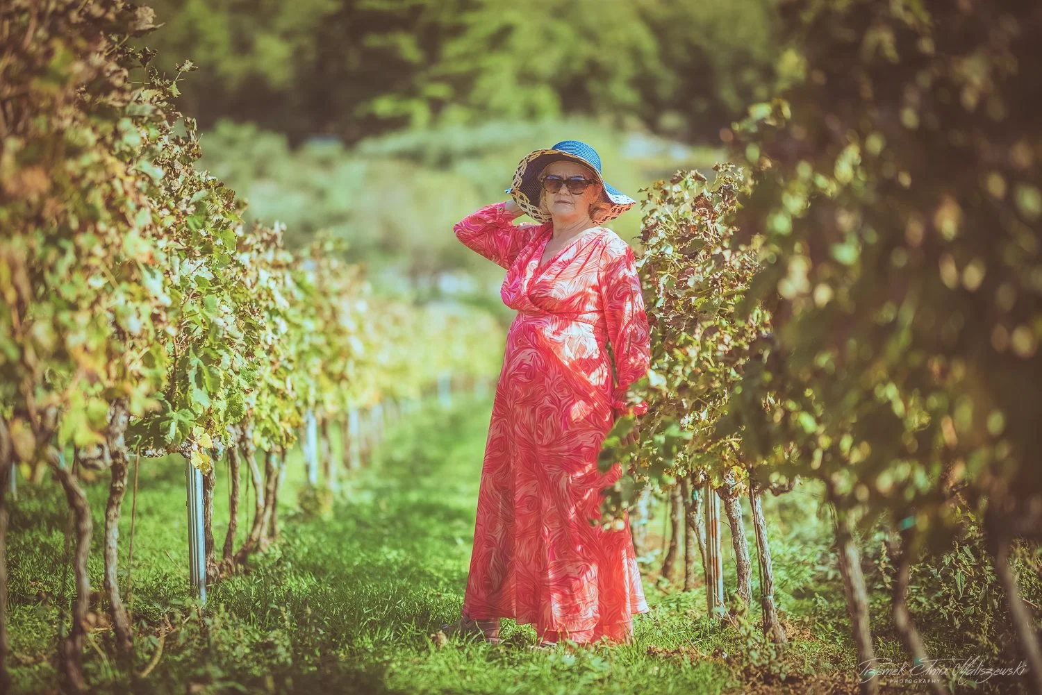 An elderly woman in a pink and orange patterned dress, wearing a large blue sun hat and sunglasses, stands in a vineyard surrounded by green foliage, holding her hat with one hand.
