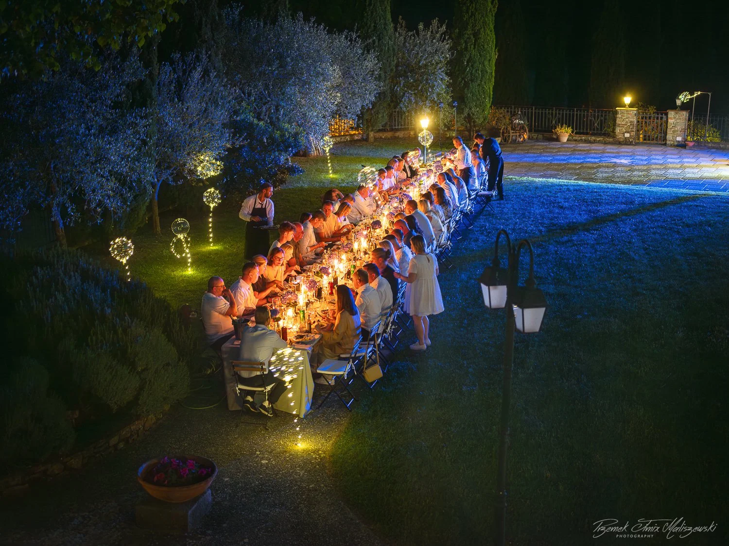 Nighttime outdoor dinner party with long table, guests seated and engaging, lit by candles and lanterns, surrounded by trees and decorative lights, with a stone patio in the background.
