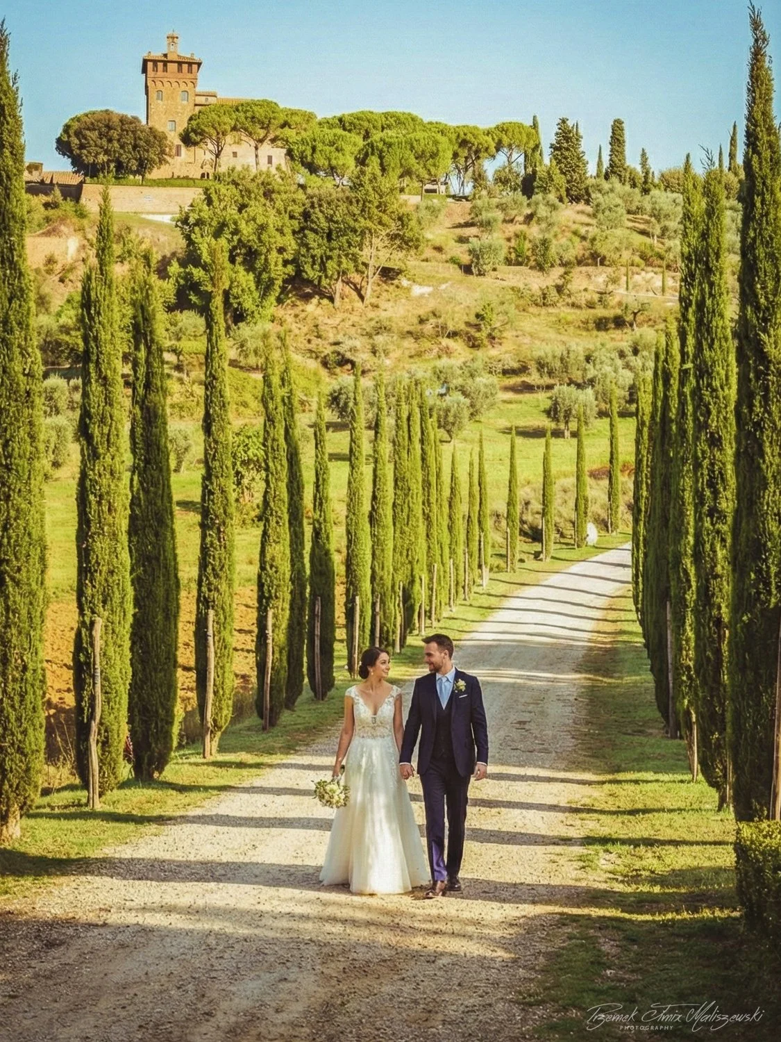 A bride and groom walking hand in hand down a tree-lined dirt path in a scenic outdoor setting, with lush green trees and a castle-like structure on a hill in the background.