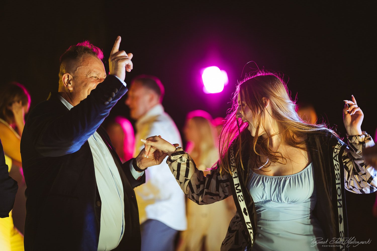 An older man and a young woman dance together at a lively party or club, holding hands and smiling. The man is wearing a dark blazer and white shirt, while the woman wears a light-colored top and a patterned jacket. Bright pink and purple lighting il