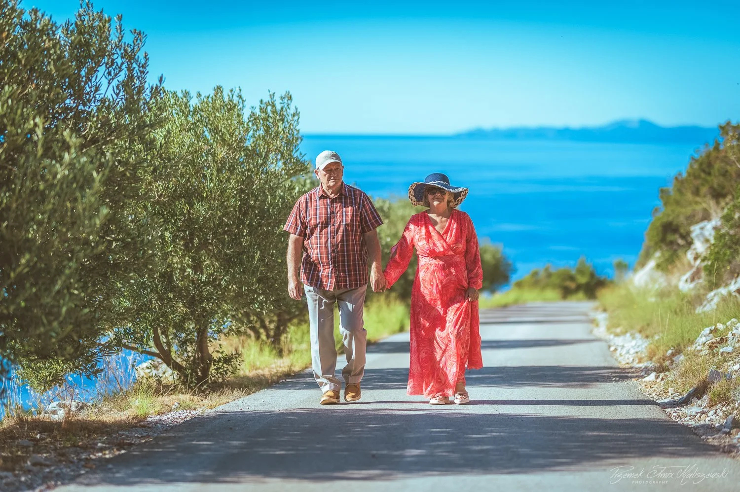 An elderly man and woman holding hands while walking along a scenic path with water and greenery in the background.