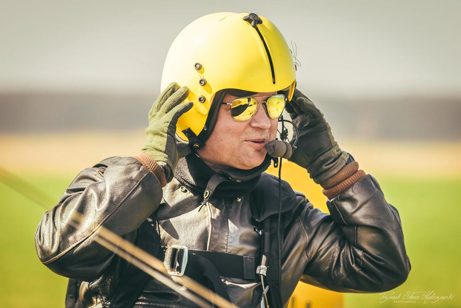 Man wearing a yellow helmet, sunglasses with reflective lenses, and a leather jacket, adjusting his helmet outdoors.
