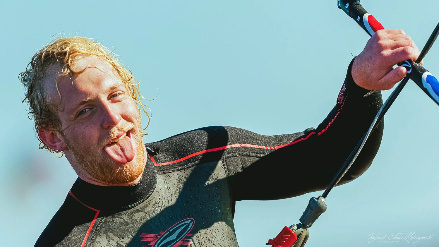 A man with blonde curly hair and a beard sticking out his tongue while holding a ski pole, wearing a wetsuit, against a clear blue sky.