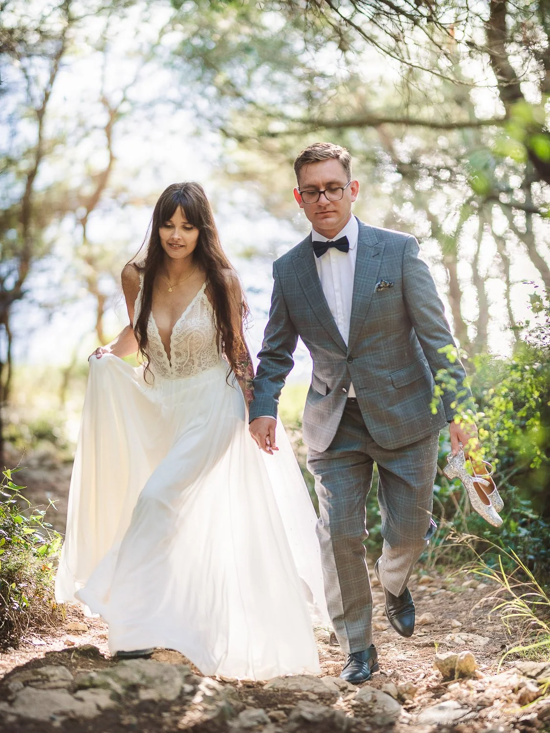 A bride and groom walking outdoors on a sunny day in a wooded area, holding hands. The bride wears a white wedding dress and the groom is dressed in a gray suit with a bow tie, holding a pair of sparkly high-heeled shoes in his right hand.
