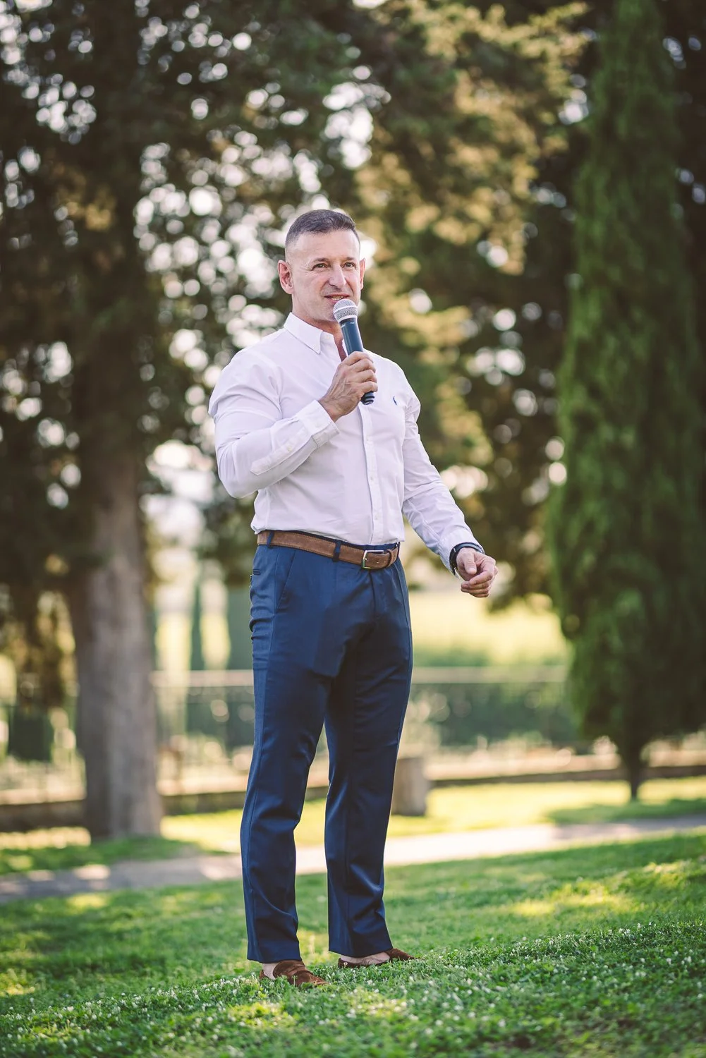 A man in a white shirt and navy pants speaking into a microphone outdoors, standing on grass with trees in the background.