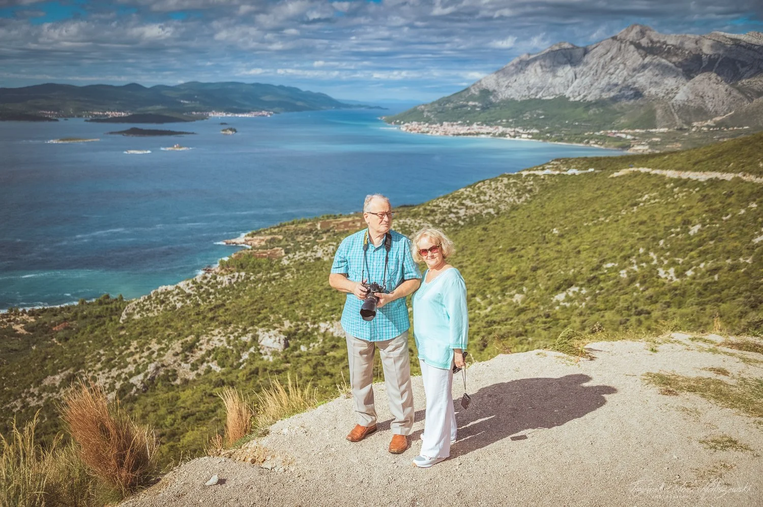 An elderly couple standing on a hill overlooking a body of water with islands, mountains, and a cloudy sky in the background. The man is holding a camera, and the woman is holding glasses.