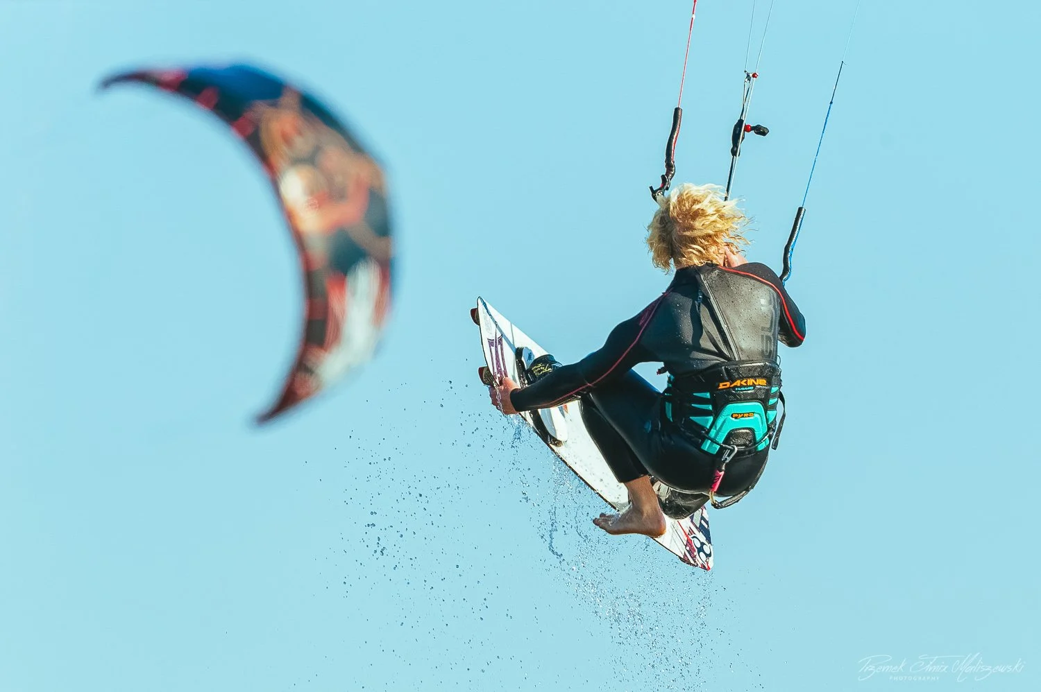 Person kiteboarding in the sky during daytime, showing a close-up of the kite in the foreground and the rider mid-air on the board.