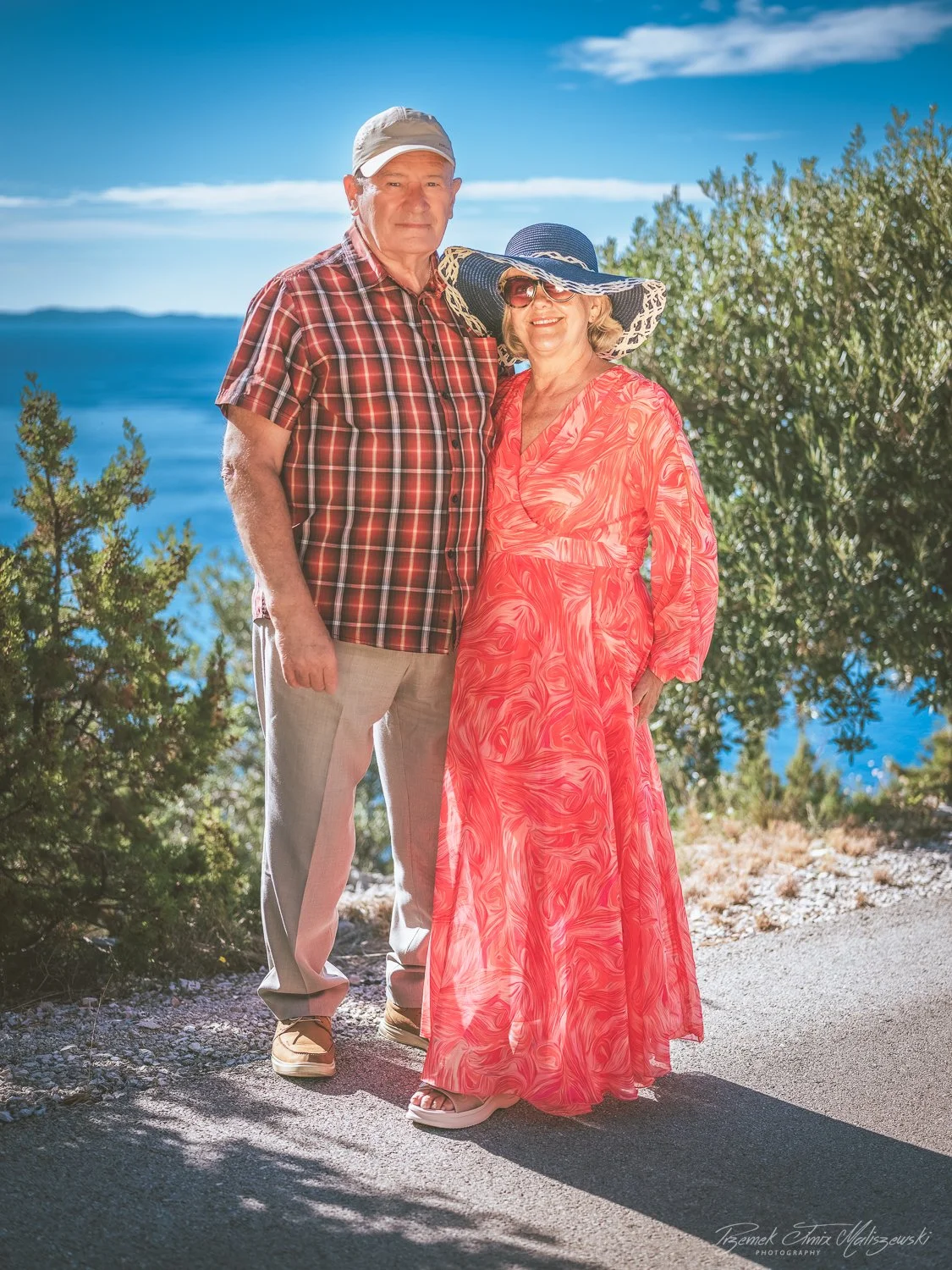 An elderly man and woman standing together outdoors by a body of water, with trees and a blue sky in the background. The man wears a plaid shirt, light pants, and a cap. The woman wears a colorful dress, large sun hat, and sunglasses.