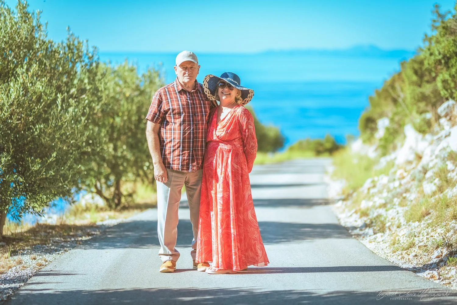 An elderly couple standing on a paved road near the water, with greenery on both sides and a bright blue sky in the background. The woman is wearing a red dress and wide-brimmed hat, and the man is in a checkered shirt and light-colored pants.