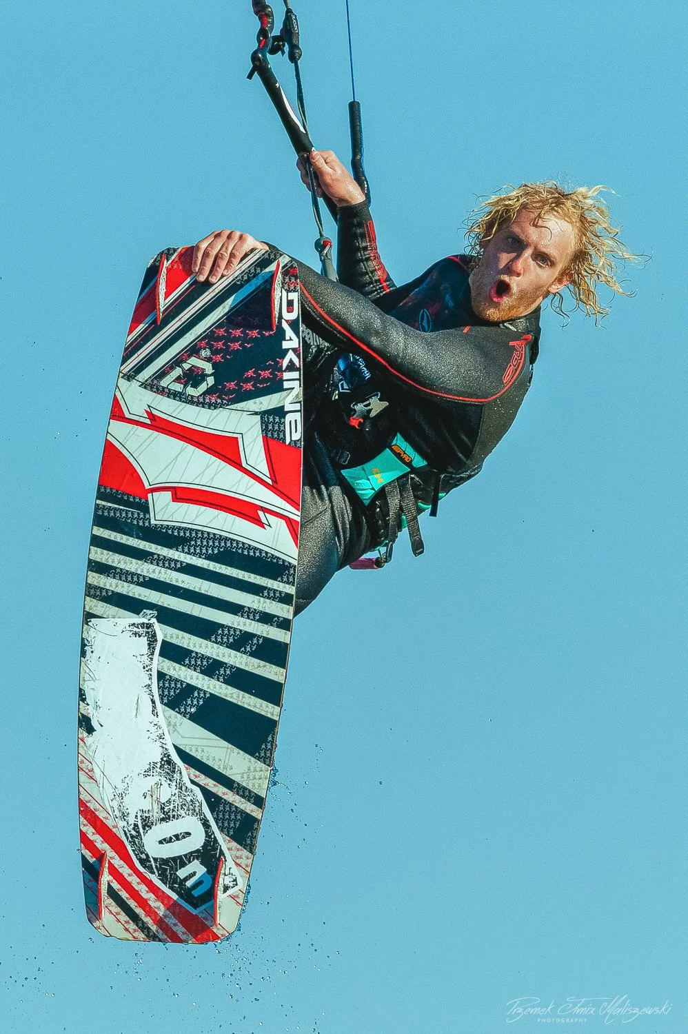 A man kiteboarding in mid-air with a colorful kiteboard against a clear blue sky.