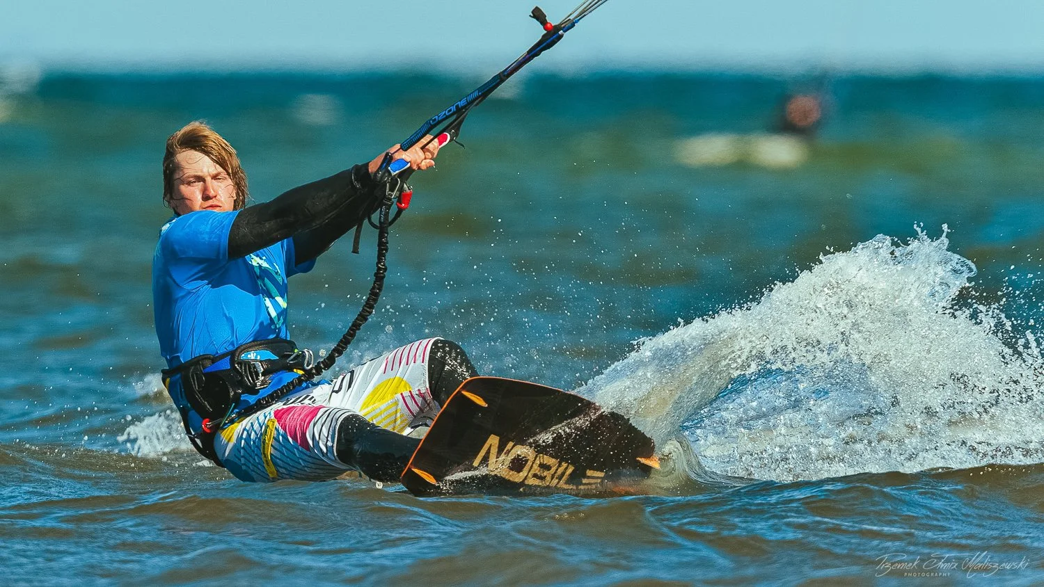 Young man windsurfing on the water, holding the sail with both hands, wearing a blue shirt, colorful shorts, and a black wetsuit sleeve on one arm, with a splash of water in the background.