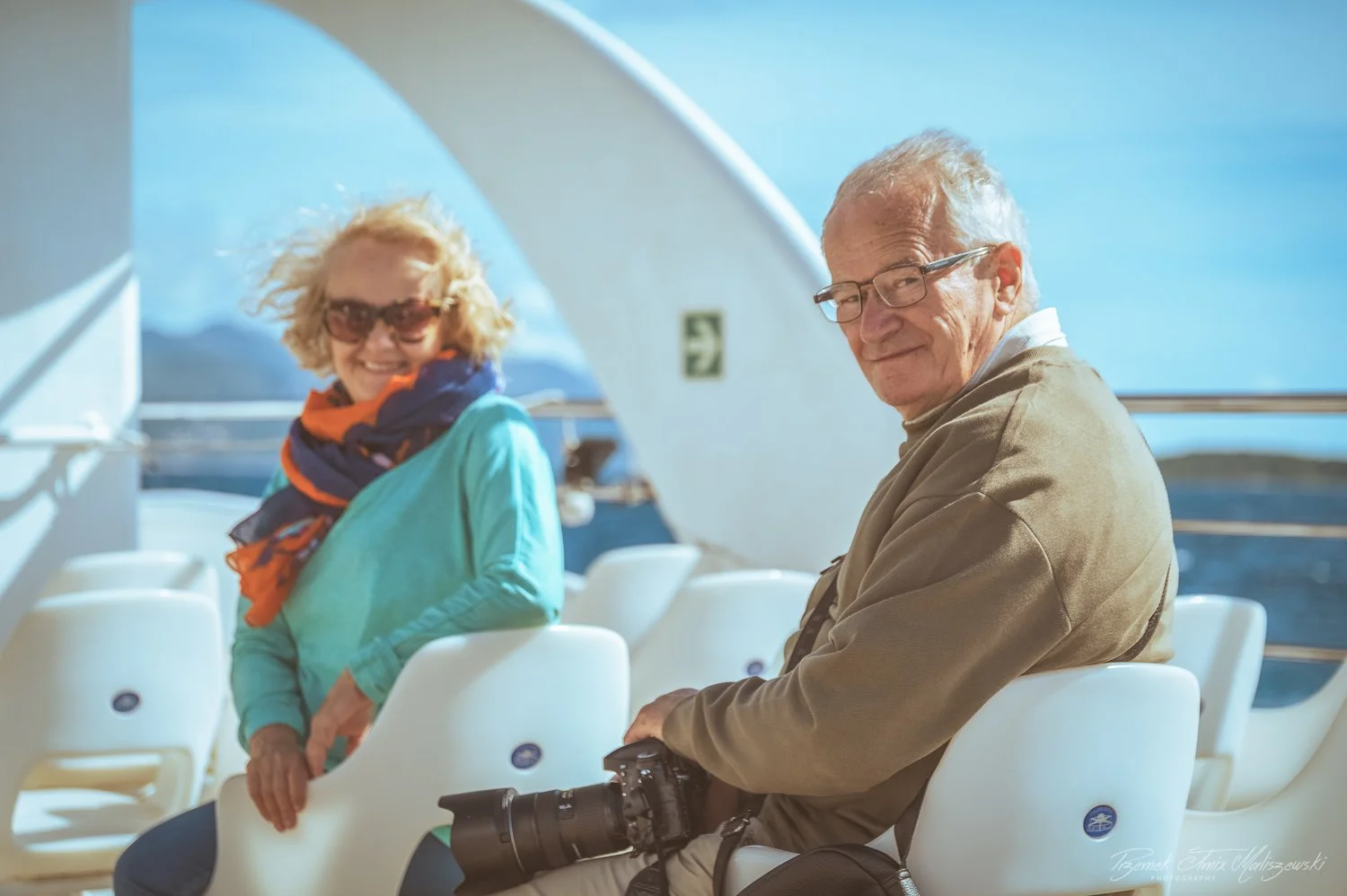 A smiling woman in sunglasses and a colorful scarf on a cruise ship with a camera and a man in glasses looking over her shoulder, with water and mountains in the background.