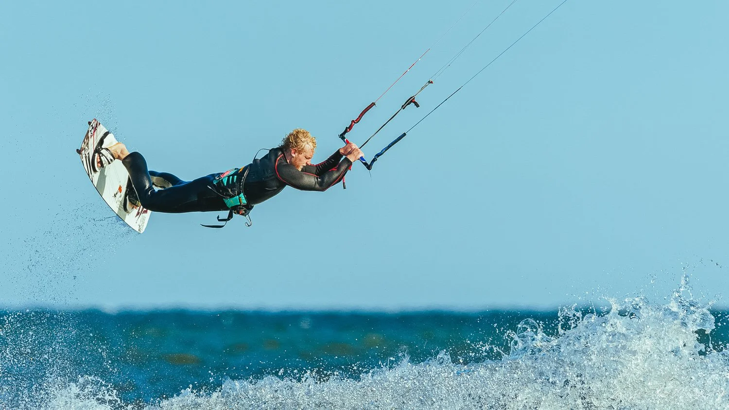 A person kiteboarding on the water, airborne above the waves with a blue sky background.