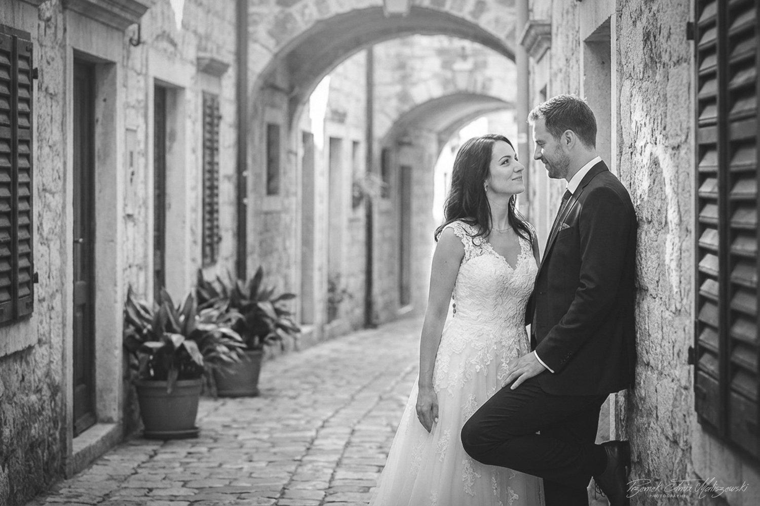 A bride and groom in wedding attire standing close, looking into each other's eyes on a cobblestone street with stone buildings and archways, black-and-white photo.