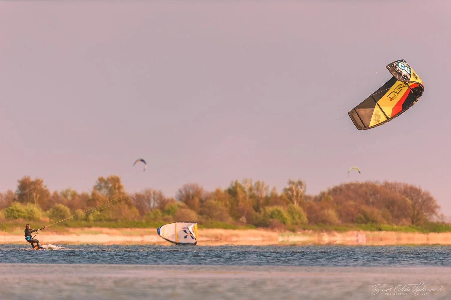 Person kite surfing on a lake with multiple kites in the sky and trees in the background.